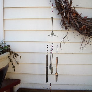 May include: A silver wind chime made of forks and knives hangs from a twig wreath. The chime is decorated with red and white beads.