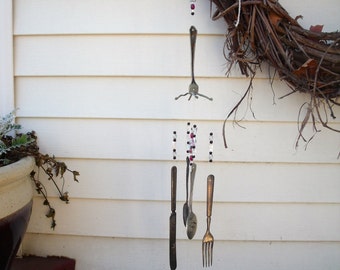 May include: A silver wind chime made of forks and knives hangs from a twig wreath. The chime is decorated with red and white beads.