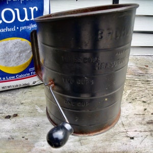 Puede incluir: Un tamizador de harina de metal negro vintage con un asa y un pomo negro. El tamizador tiene medidas para una, dos y tres tazas. Una bolsa de harina está en el fondo. La bolsa dice "Flour" y "Peso neto 2,26 kg".