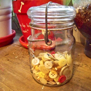 Op de afbeelding: Een glazen pot gevuld met een verscheidenheid aan witte en gele knopen. De pot heeft een metalen deksel en een draadhandvat.