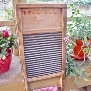 Puede incluir: Un antiguo lavadero de madera con una superficie corrugada de metal. El lavadero tiene un pomo rojo y la palabra "Washboard" escrita en la parte superior. El lavadero está sentado sobre una superficie de madera con una planta en maceta roja en primer plano.