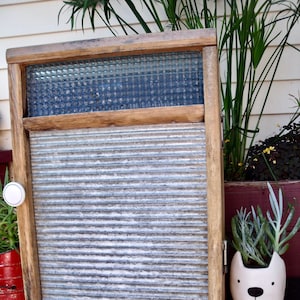 May include: A vintage wooden washboard with a metal corrugated surface and a glass window pane. The washboard is mounted on a wooden frame and has a white knob on the side. A small potted plant is in the foreground.