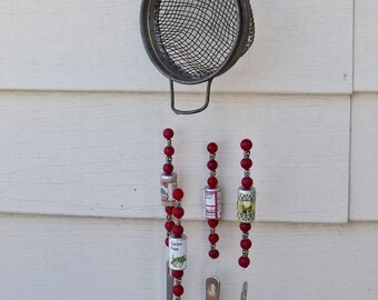 May include: A red and white kitchen utensil wind chime with a strainer, spoons, and small jars with red beads. The wind chime is hanging from a white wall.