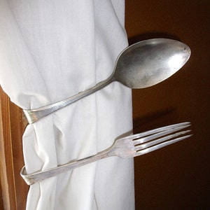 May include: A white curtain with a silver spoon and fork used as a curtain tieback.