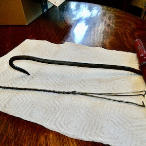 May include: A vintage metal hook with a wooden handle and a wire hook. The hook is on a white paper towel on a wooden surface.