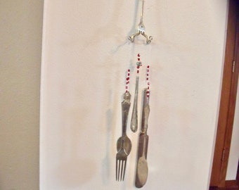 May include: A silver wind chime made from forks and spoons hanging from a white wall. The chime is decorated with red and white beads.