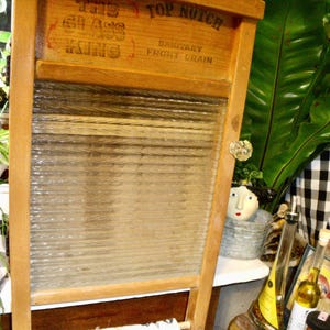 以下が含まれることがあります： ガラス製の研磨面を備えた、ヴィンテージの木製洗濯板。「THE GLASS KINGS」と「TOP NOTCH SANITARY FRONT DRAIN」の文字が上部に表示されています。洗濯板は明るい茶色の木材で縁取られています。