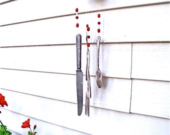 May include: A metal wind chime made from silverware and a metal bowl. The wind chime is hanging from a white wall and has red and white beads.