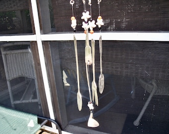 May include: A silver wind chime with five spoons and white ceramic flowers hanging from a string. The chime is hanging from a window frame.
