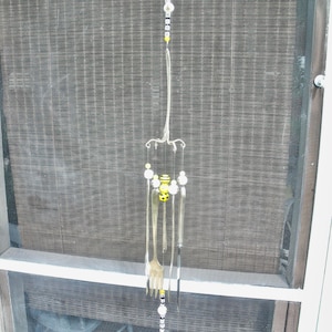 May include: A silver wind chime made of spoons and forks with black and yellow beads hanging from it. The chime is hanging from a window screen.