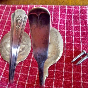May include: Two silver spoons with ornate handles have been repurposed into wall hooks. The spoons are mounted to a red and white checkered surface with screws.
