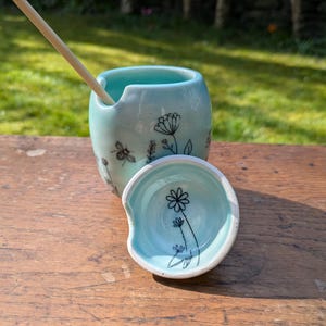 Peut inclure: Un pot à miel en céramique bleu clair avec un couvercle assorti, décoré d'illustrations florales et d'abeilles noires. Un dipper à miel en bois est à l'intérieur du pot. Le pot et le couvercle sont sur une surface en bois.