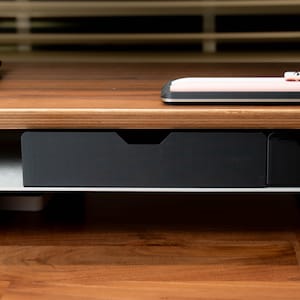 May include: A black drawer with a silver metal frame sits below a wooden desk surface. The drawer is part of a desk organizer.