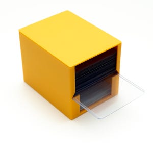 May include: A bright yellow, cube-shaped card holder with a clear plastic card protector extending from the front. The holder is filled with dark cards, creating a contrast with the yellow exterior. The design is simple and modern.