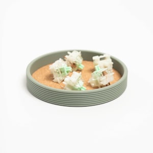 May include: A close-up of five clear, white mechanical keyboard keycaps with green accents, arranged on a cork coaster inside a light green silicone tray.