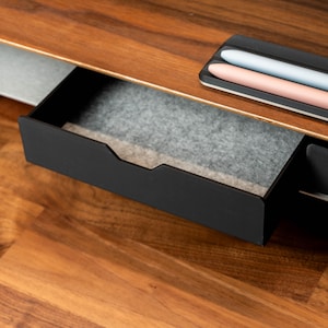 May include: A black drawer with a gray felt lining is open, revealing the interior of the drawer. The drawer is part of a wooden desk with a light brown finish.