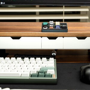 May include: A wooden desk organizer with two white drawers and a black tray for holding small items. The tray contains a collection of colorful keycaps. A white keyboard and a black mouse are on the desk.