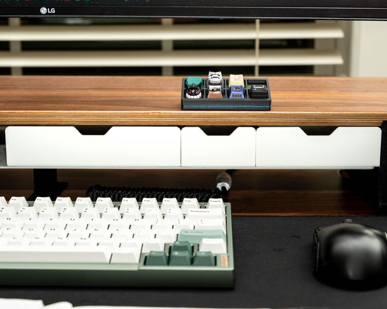 May include: A wooden desk organizer with two white drawers and a black tray for holding small items. The tray contains a collection of colorful keycaps. A white keyboard and a black mouse are on the desk.