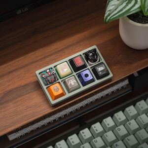 May include: A green tray with a variety of colorful keycaps. The keycaps feature different designs, including a skull, a robot, and Japanese characters. The tray is sitting on a wooden surface.