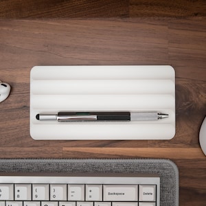 Pode incluir: Um porta-canetas branco com três ranhuras para canetas ou lápis. Uma caneta prateada com cabo preto está apoiada na ranhura superior. O porta-canetas está sobre uma superfície de madeira.