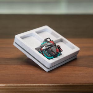 以下が含まれることがあります： グレー、赤、ターコイズの小さな詳細なロボットの頭部の置物が、3つの区画のある白い斑点のあるプラスチック製のディスプレイトレイに入っています。