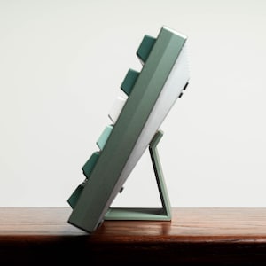 May include: A green mechanical keyboard with white and teal keycaps, propped up on a matching stand. The keyboard is angled, showcasing its side profile and key arrangement. The stand provides support, creating an ergonomic typing angle.