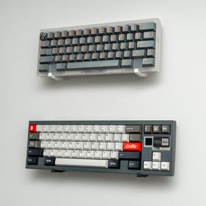 May include: Two keyboards mounted on a white wall. The top keyboard is a 60% keyboard with a dark gray case and orange keycaps. The bottom keyboard is a full-sized keyboard with a dark gray case and white keycaps. The bottom keyboard has a red keycap with the text "Creamsicle" printed on it.