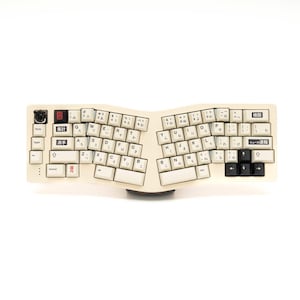 Könnte beinhalten: Eine weiße geteilte Tastatur mit japanischen Zeichen auf den Tasten. Die Tastatur hat ein Schwarz-Weiß-Farbschema und ist ergonomisch gestaltet.