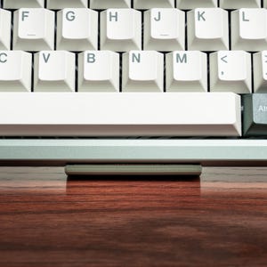 May include: Close-up of a white mechanical keyboard with a silver base. The keys are white with black lettering. The space bar is long and white. A small, rectangular device rests on a wooden surface in front of the keyboard.