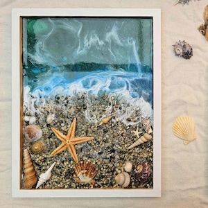 May include: A framed resin art piece depicting a beach scene with blue and green water, white waves, and brown pebbles. The artwork includes a starfish, seashells, and other beach elements.