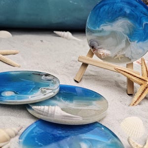May include: Four round resin coasters with a blue and white ocean wave design. The coasters are on a white sandy surface with seashells and a starfish.