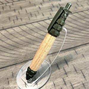 Puede incluir: Un bolígrafo de madera y metal verde con forma de granada. El bolígrafo está en un soporte de plástico transparente.