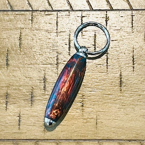 May include: A handcrafted keychain with a vibrant, swirled design in shades of red, blue, and brown. The keychain features a polished, cylindrical body and a silver-colored metal ring for attaching keys. The overall length is approximately 4 inches.