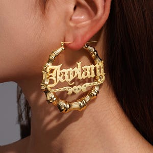 May include: Gold-colored bamboo-style hoop earrings. The earrings feature the name "Jaylan" in a stylized font, with a heart design. The earrings are worn on a person's ear.