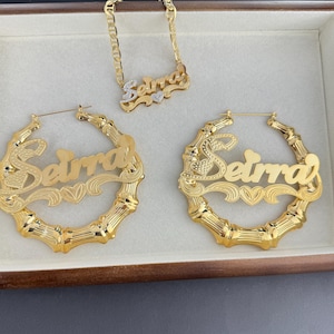 May include: A gold-colored jewelry set featuring a necklace and hoop earrings, all with the name "Seirra". The earrings have a bamboo design. The necklace and earrings are displayed in a wooden box.