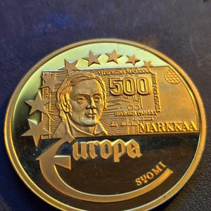 May include: A gold-colored coin featuring a portrait of a person, the word "Europa", and the text "500 Markkaa". The coin has a star design and the text "Suomi" on the bottom.