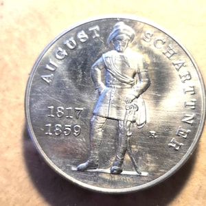 May include: A silver coin featuring a detailed relief of a man in historical attire, with the inscription "AUGUST SCHARTNER" and the dates "1817 1859" encircling the figure. The coin has a worn, antique appearance.