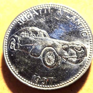 May include: A silver coin featuring a detailed engraving of a Bugatti Atlantic car. The coin is inscribed with "BUGATTI ATLANTIC" and the year "1937". The coin has a detailed edge and a slightly worn appearance.