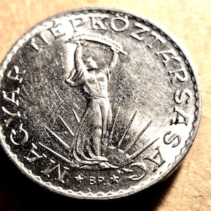May include: A silver coin featuring a woman holding a sheaf of wheat, surrounded by text. The coin has a detailed design with radiating lines. The text includes the words "NÉPKÖZTÁRSASÁG" and "BP."