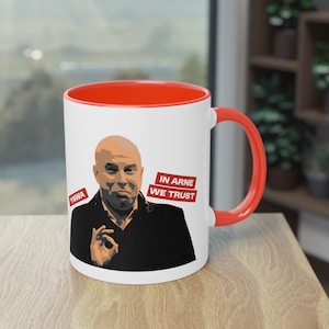 Op de afbeelding: Witte keramische mok met een rode rand en handvat. De mok heeft een cartoonillustratie van een man met de tekst "IN ARNE WE TRUST" en "YNWA" op de mok.