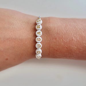 May include: White beaded bracelet with gold lettering spelling out "COUGARS".