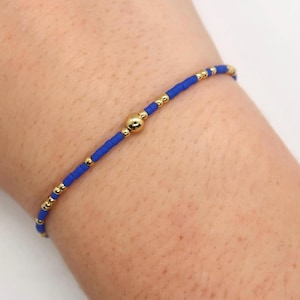 May include: A blue and gold beaded bracelet with a gold bead accent. The bracelet is on a person's wrist.