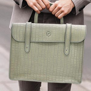 May include: A light green briefcase with a textured, crocodile-like pattern. The briefcase has a top handle, a flap closure, and two vertical straps. The bag is held by a person wearing a gray pinstripe suit.