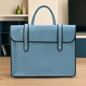 May include: A light blue briefcase with a textured, crocodile-like pattern. The bag has a top handle and two straps with gold-colored rivets. The edges are trimmed with black. The bag is sitting on a wooden surface.
