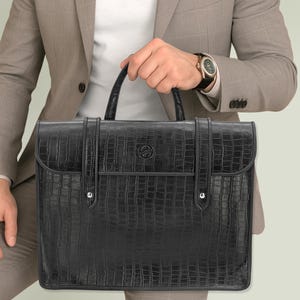 Op de afbeelding: Zwarte aktetas met een krokodillenleertextuur, met een handvat aan de bovenkant en een klepsluiting. De tas heeft zilverkleurige hardware en een klein embleem aan de voorkant. De aktetas wordt vastgehouden door een persoon in een pak.