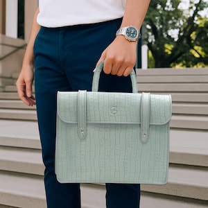 Op de afbeelding: Een lichtgroene aktetas met een textuurpatroon dat op krokodillenleer lijkt. De aktetas heeft een handvat aan de bovenkant, een klepsluiting en twee riemen. De tas wordt vastgehouden door een persoon die een wit overhemd en een marineblauwe broek draagt.