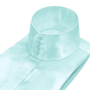 Puede incluir: Una camisa azul claro con cuello alto y redondeado y cuatro pequeños botones transparentes. La camisa está hecha de un material liso, similar al satén. El cuello está abotonado.