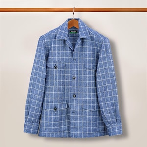 Puede incluir: Chaqueta a cuadros azul y blanco con mangas largas, dos bolsillos en el pecho y dos bolsillos inferiores. La chaqueta tiene una parte delantera abotonada y está colgada de una percha de madera.