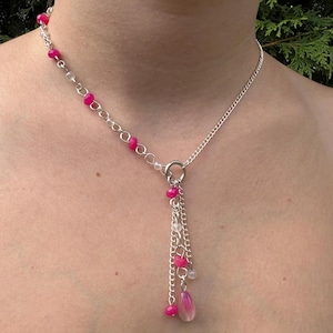 May include: A silver necklace with a delicate design. The necklace features a chain with pink and clear beads, and a dangling cluster of chains with pink beads and a pink teardrop-shaped charm. The necklace is worn around the neck.