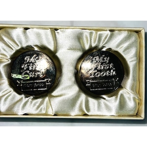 Könnte beinhalten: Zwei silberfarbene Andenkenmünzen, eine mit der Aufschrift "My First Curl" und die andere "My First Tooth", präsentiert in einer satin gefütterten Box. Die Münzen haben eine Vintage-Ästhetik, perfekt für die Aufbewahrung von Babyerinnerungen.